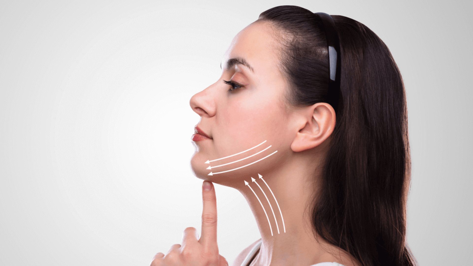 Profile of a woman with long dark hair, wearing a headband. White arrows on her jawline indicate a lifting motion, suggesting a concept of facial treatment.