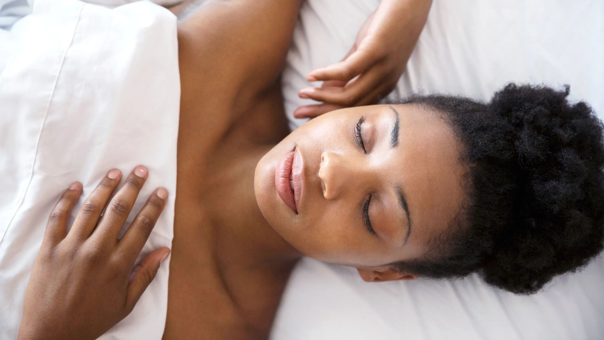 A person peacefully sleeping on a white bed with relaxed facial expressions, their hand resting on the sheet. The scene conveys a sense of calm and restfulness.