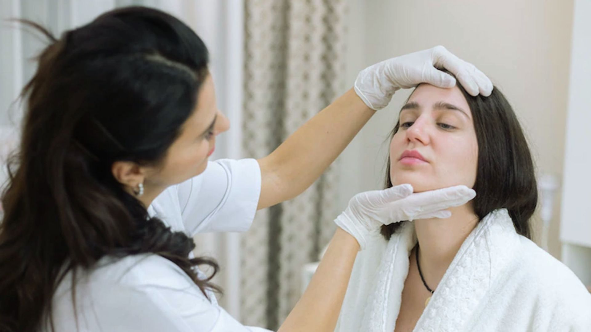 A dermatologist wearing gloves examines a woman's face in a clinical setting. The woman appears relaxed, highlighting a professional skincare consultation.