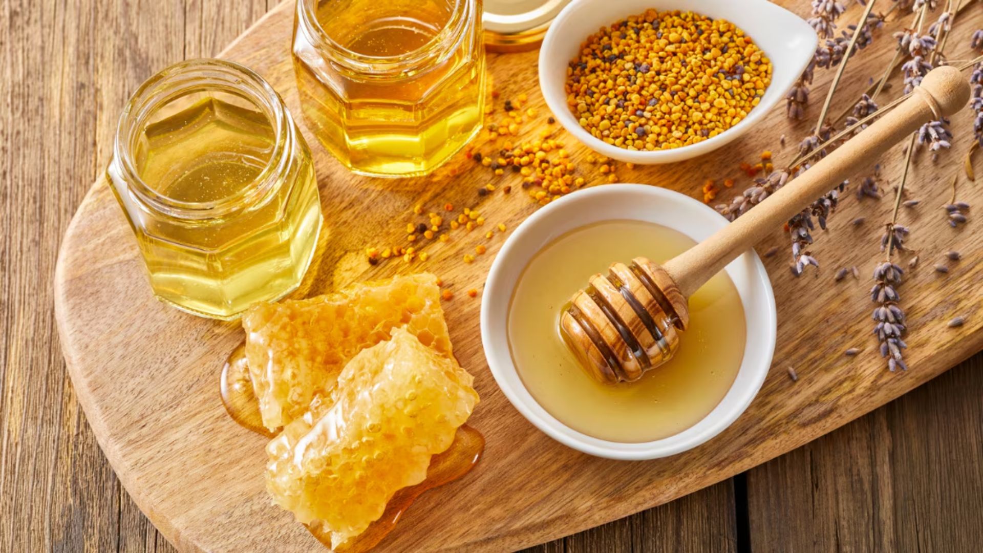 Wooden board with jars of honey, honeycomb, pollen in white bowls, and lavender sprigs. A honey dipper rests in a small dish, creating a rustic and natural feel.