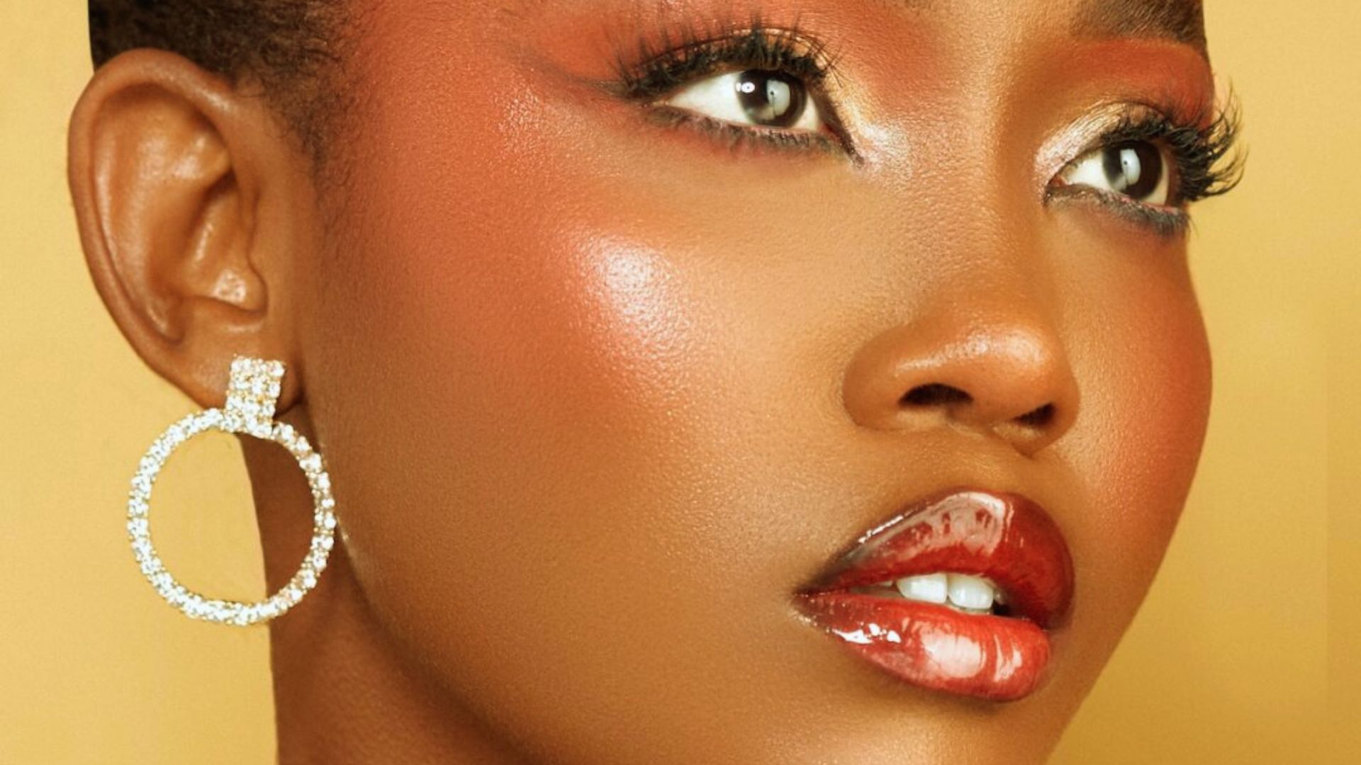 Portrait of a woman with glowing bronze skin, glossy red lips, glittery eyeshadow, and sparkling hoop earrings against a warm, golden background.