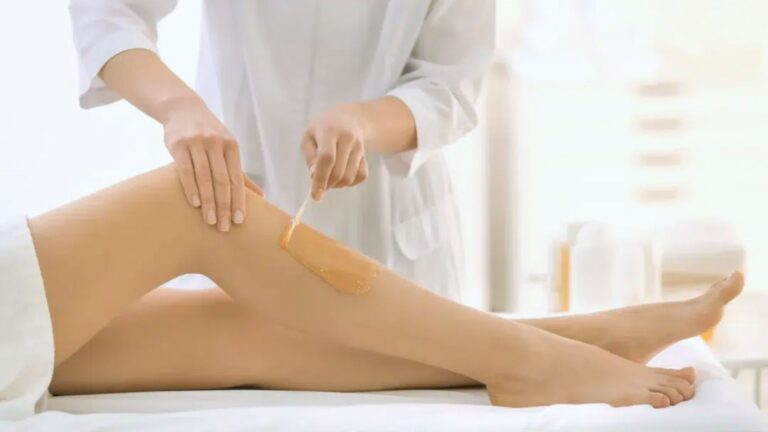 An aesthetician in a white coat applies wax to a woman's lower leg in a bright, clean spa setting, conveying a calm and professional atmosphere.