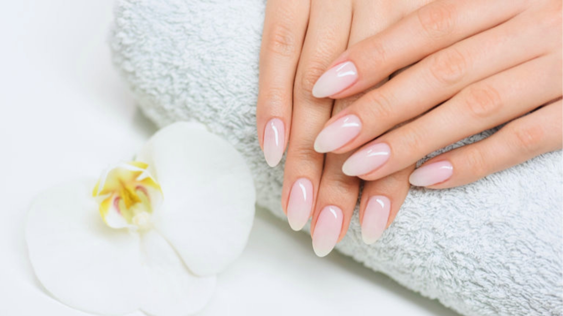 Hands with manicured nails in a soft pink ombre style resting on a white towel, next to a delicate white orchid, conveying elegance and calm.