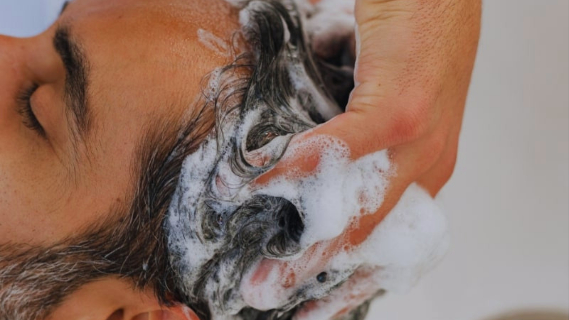 Close-up of a person having their hair washed, with hands massaging shampoo into the scalp. The scene conveys a sense of relaxation and cleanliness.