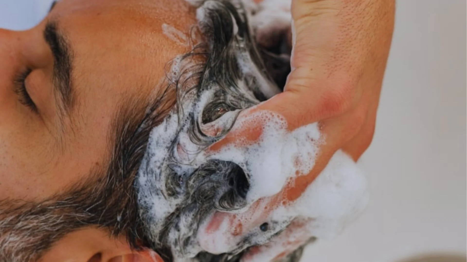 Close-up of a person having their hair washed, with hands massaging shampoo into the scalp. The scene conveys a sense of relaxation and cleanliness.