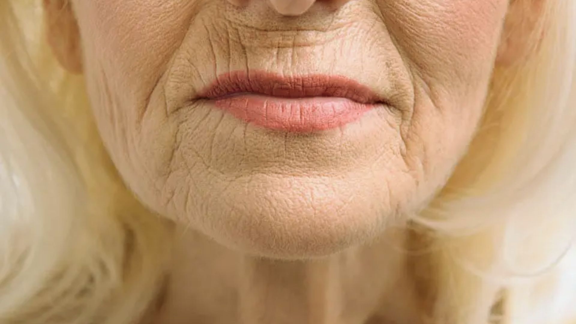 Close-up of an older woman's mouth, showing lips and surrounding skin with deep wrinkles. The expression is calm; her light hair frames the image edges.