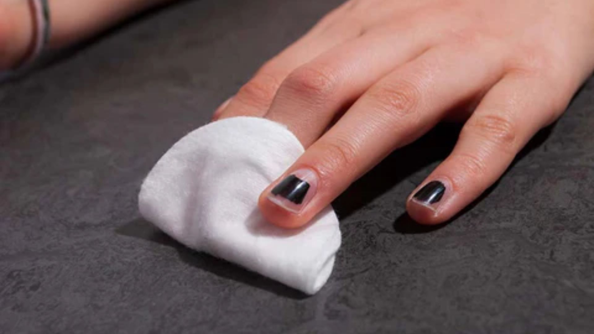 A hand with black nail polish gently rests on a dark marble surface. The index finger is partially covered by a white cotton pad, suggesting nail care.