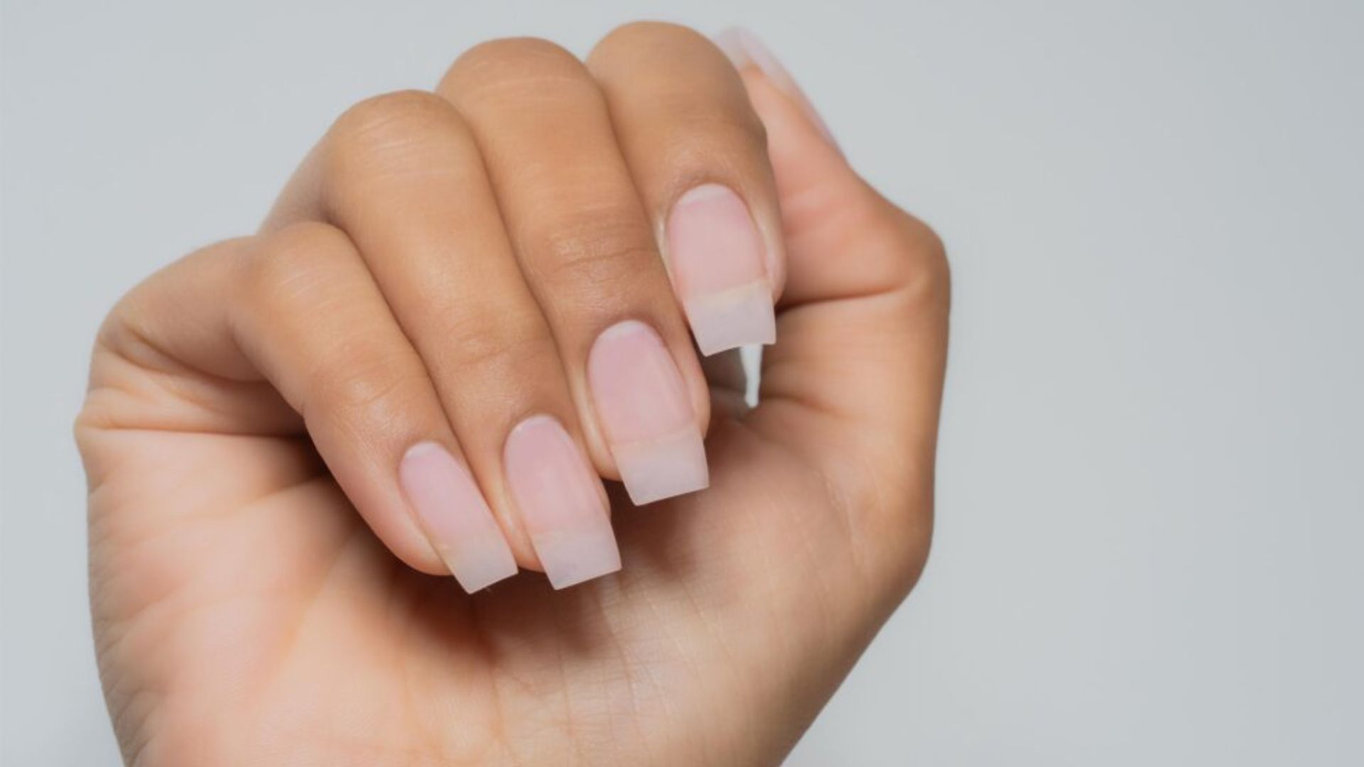 A hand with a light skin tone displays clear, square-shaped acrylic nails against a plain gray background, conveying elegance and simplicity.
