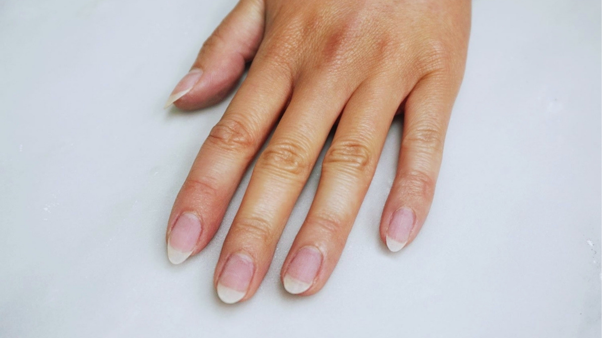 A hand with natural, unpolished nails rests on a light surface. The nails are long, shaped as a soft point. The skin tone is light, conveying simplicity.