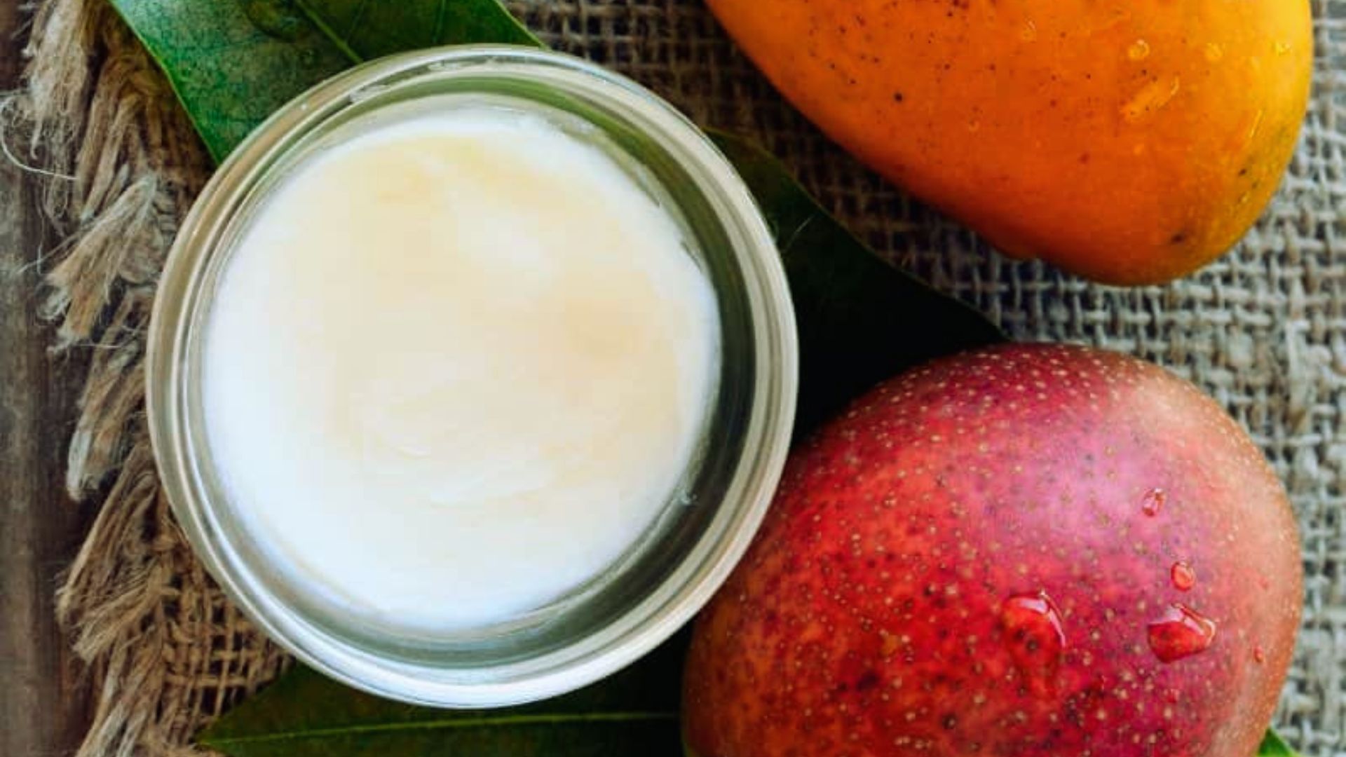 A jar of creamy white lotion sits on a green leaf alongside a ripe orange and red mango on rustic burlap, creating a fresh, tropical vibe.