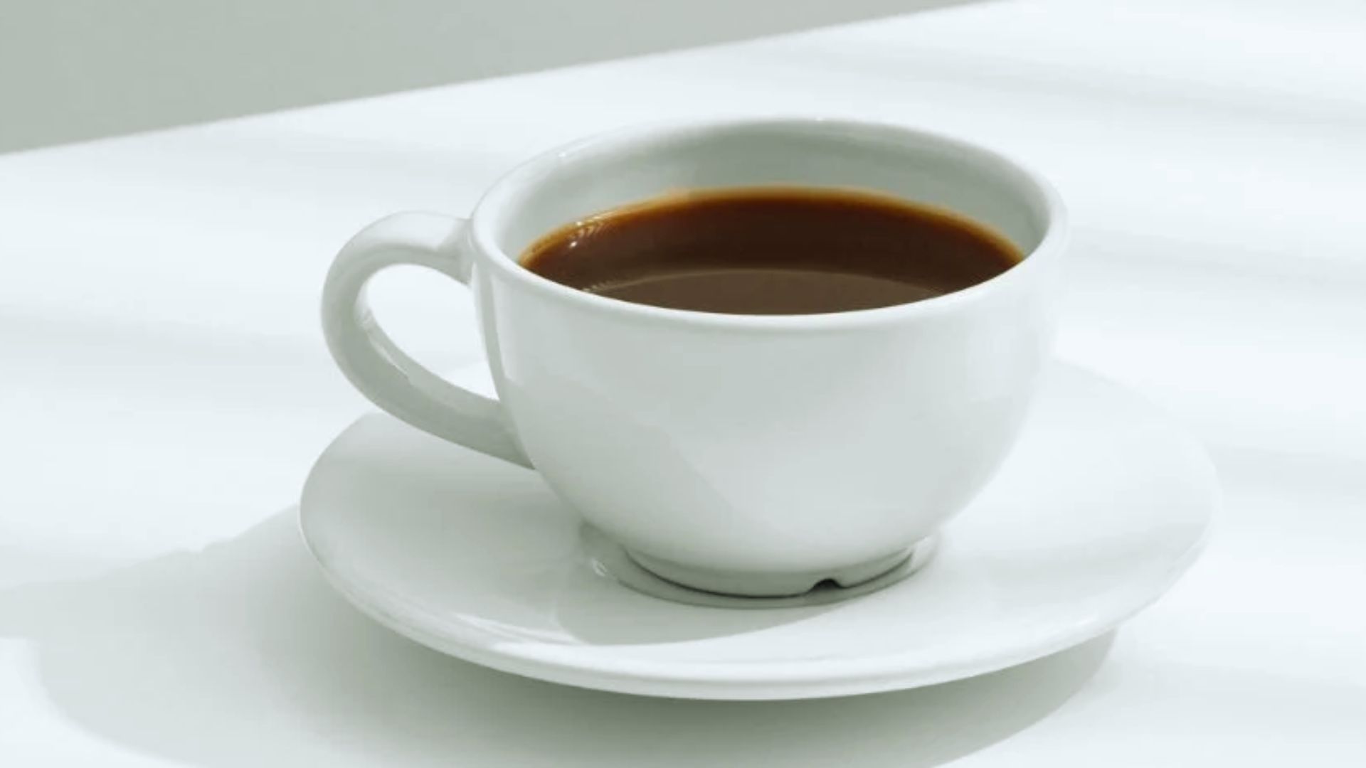 A white cup filled with dark coffee sits on a matching saucer against a light-colored background, conveying simplicity and calm.