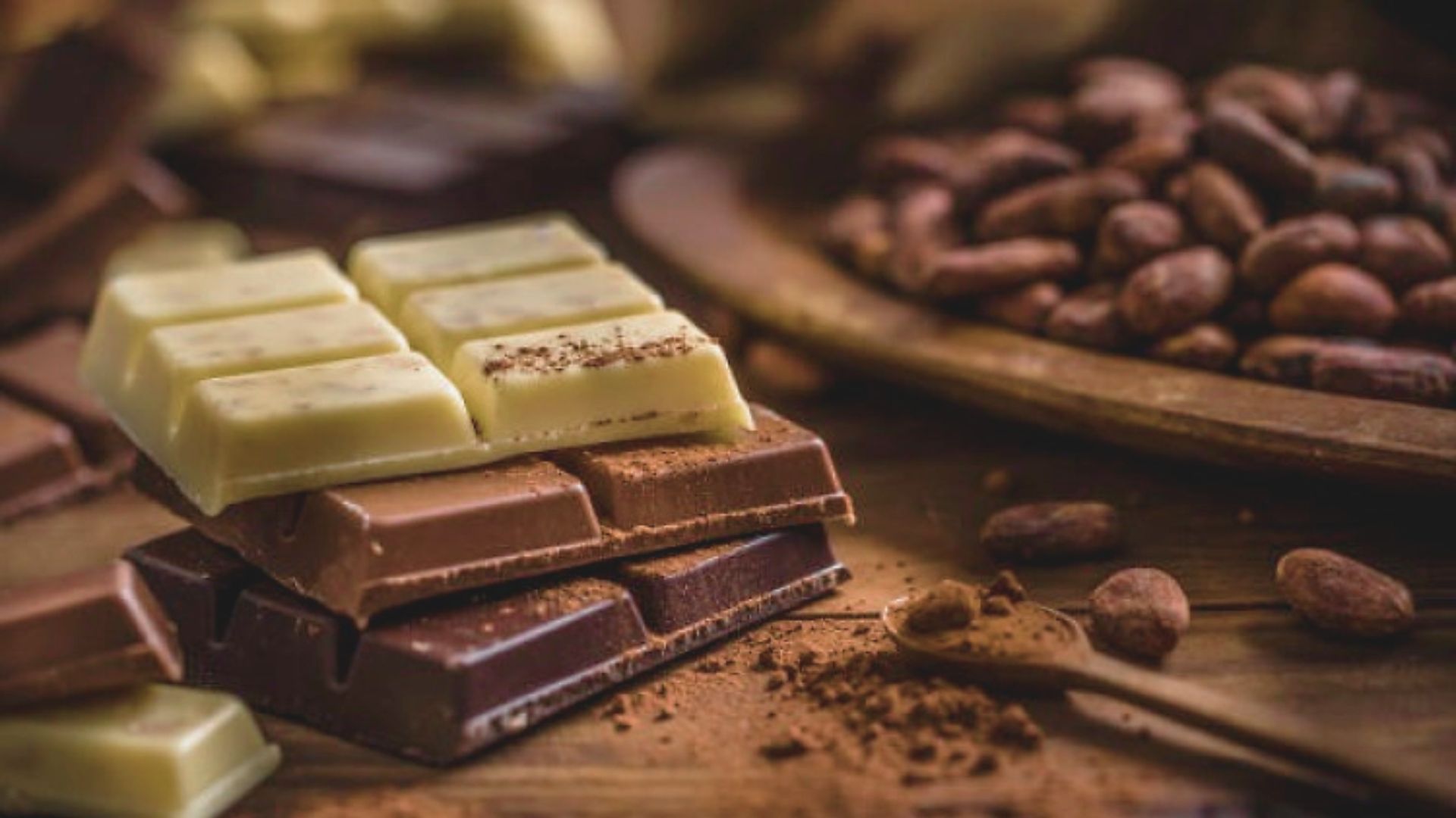 Assortment of chocolate bars including white, milk, and dark chocolate stacked on a rustic table with cocoa beans and powder, creating a rich, indulgent mood.