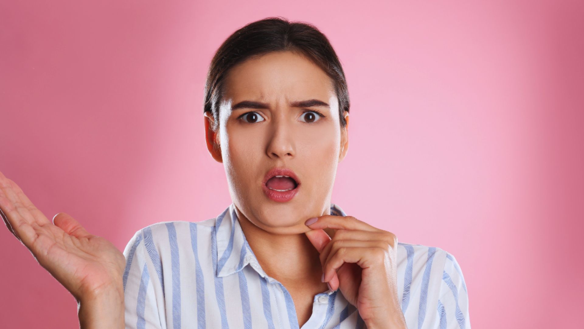 Woman with a surprised expression, wearing a striped shirt, gestures with open palms against a pink background, conveying shock or confusion.