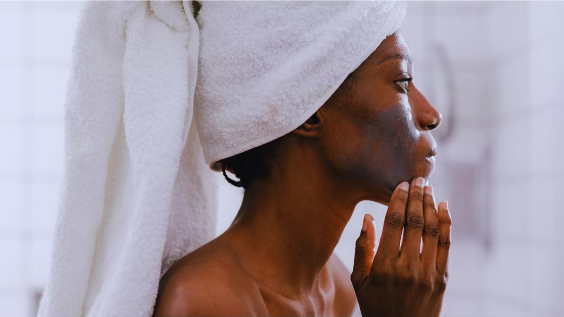 A person with a towel wrapped around their head applies a clay face mask in a bathroom. The setting conveys a calm, self-care routine.