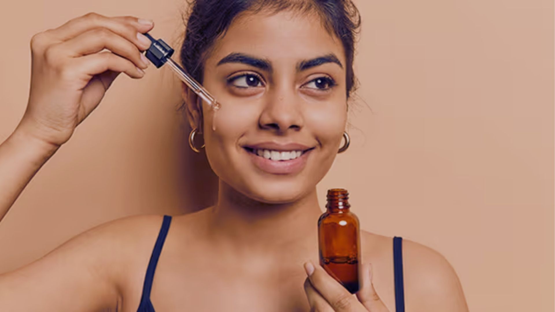 Smiling woman applies face serum with dropper, holding a brown bottle. She stands against a beige background, conveying a sense of skincare routine.
