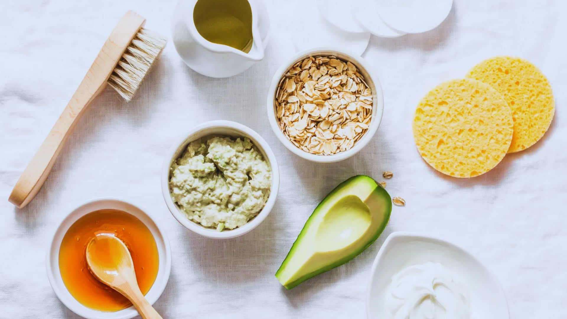 A flat lay of DIY spa ingredients on a white surface, including avocado, oats, honey, a wooden brush, yellow sponges, olive oil, and cream, conveying a serene, natural spa vibe.