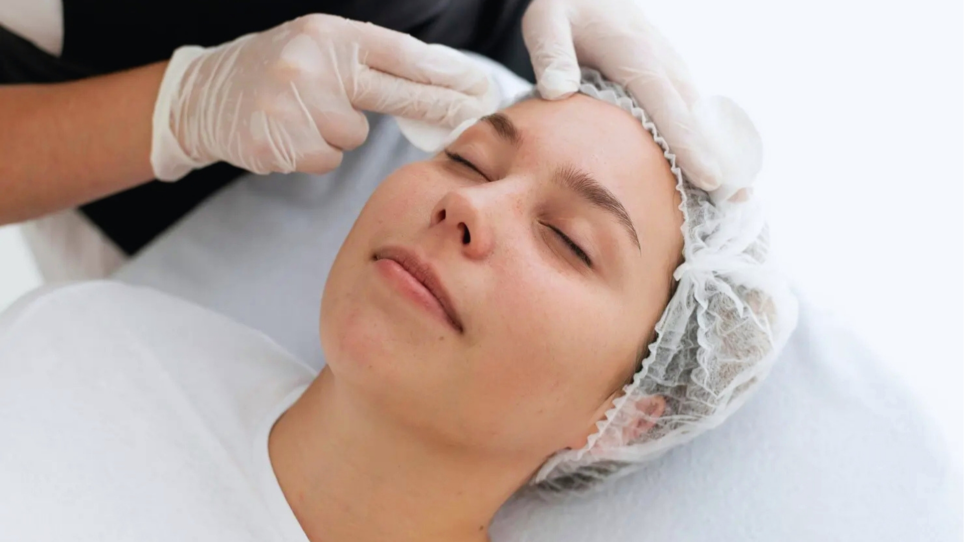 A person receives a facial treatment, eyes closed and relaxed. They're wearing a hair cap, while hands in gloves gently apply care to their forehead. Calm and serene atmosphere.