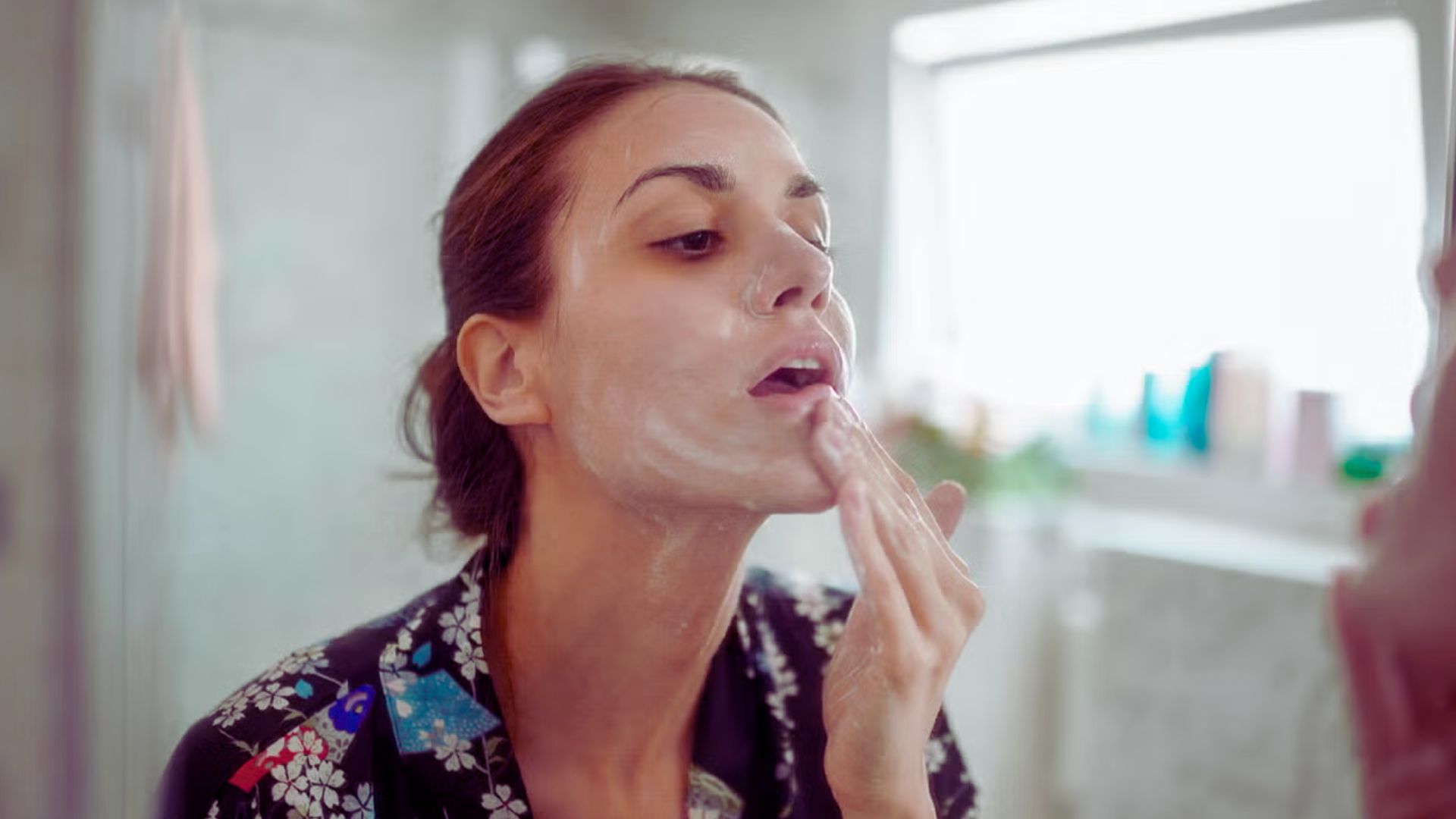 Woman applying a creamy face mask in a bathroom, looking at her reflection. The atmosphere feels calm and focused, with soft lighting and a floral robe.