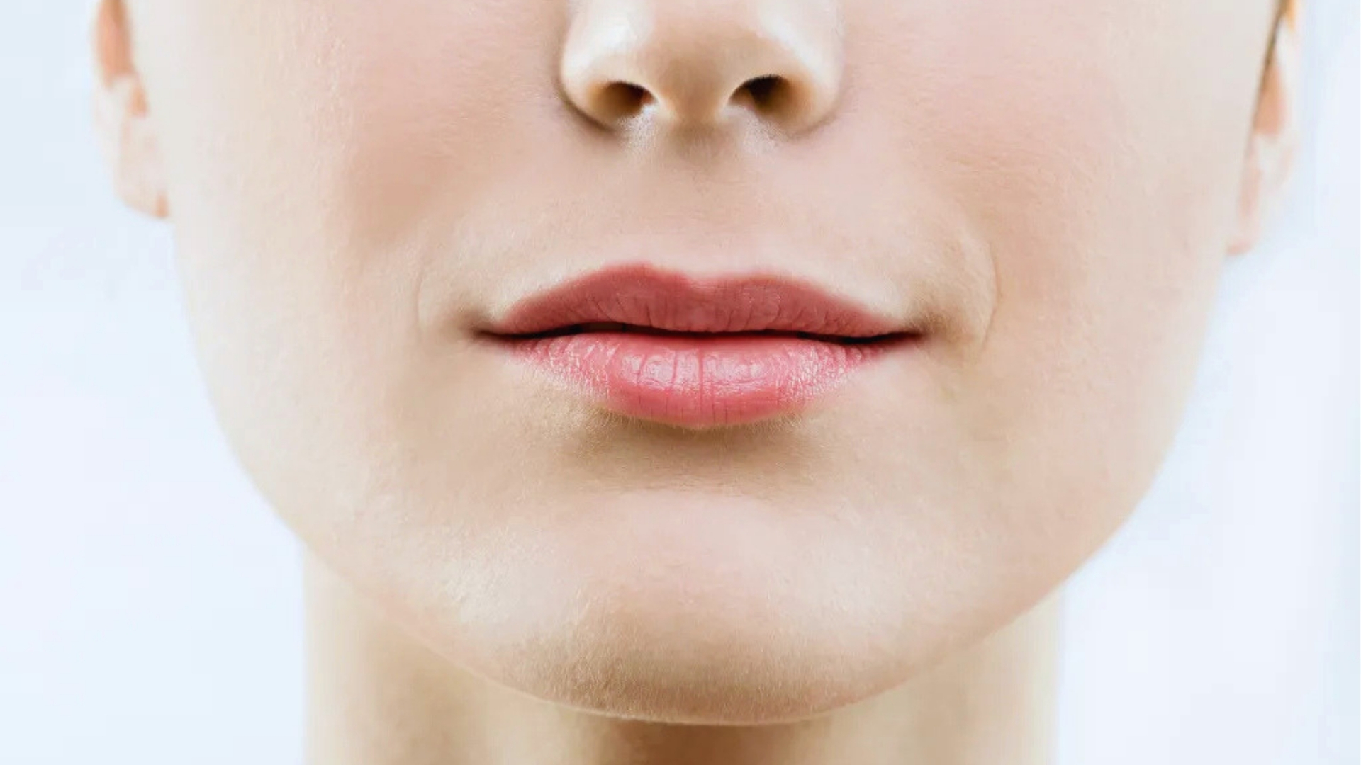 Close-up of a person's lips and chin, showing smooth skin and slightly parted lips with a neutral expression. The background is softly blurred and light.