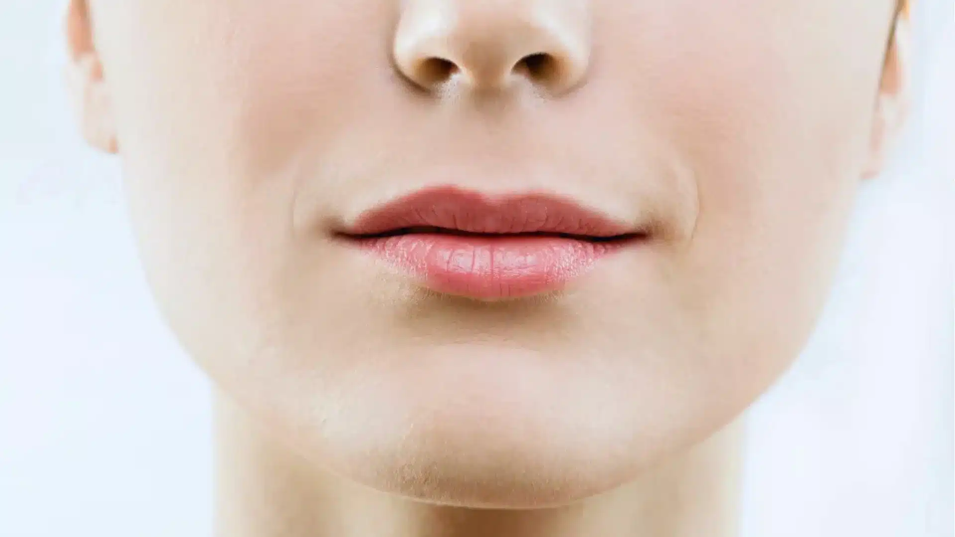Close-up of a person's lips and chin, showing smooth skin and slightly parted lips with a neutral expression. The background is softly blurred and light.