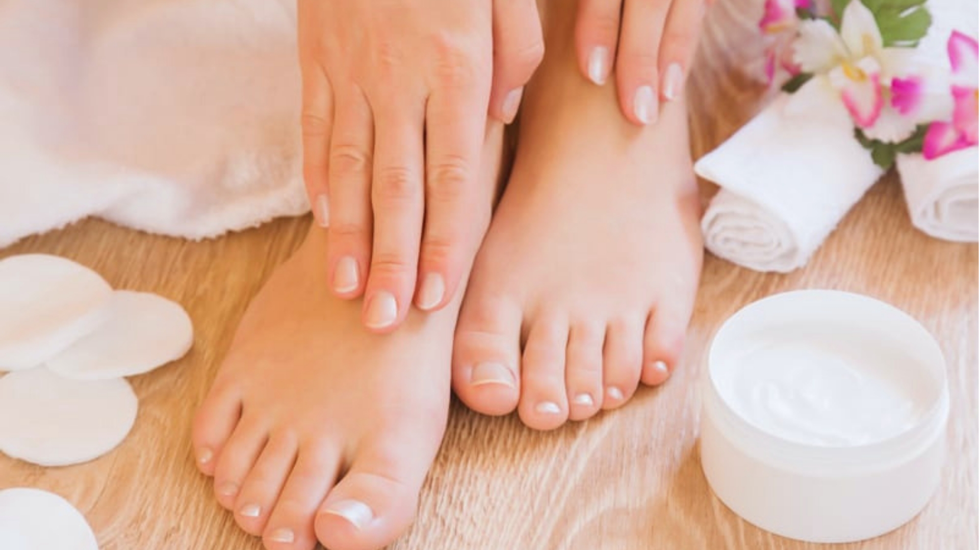Hands gently touch feet on a wooden floor, surrounded by beauty items like cotton pads, a jar of cream, and fresh flowers, creating a serene spa vibe.