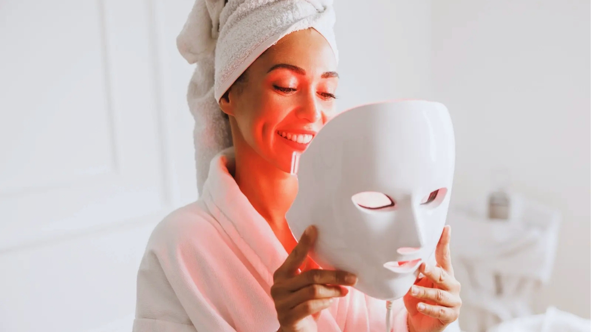 A woman in a bathrobe and towel on her head smiles while holding a glowing LED face mask, conveying relaxation and skincare routine in a spa-like setting.