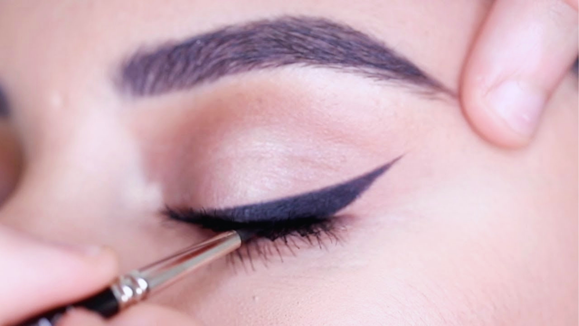Close-up of an eyelid with winged eyeliner being applied using a brush. The eye is gently closed, showing precise makeup application and defined eyebrows.