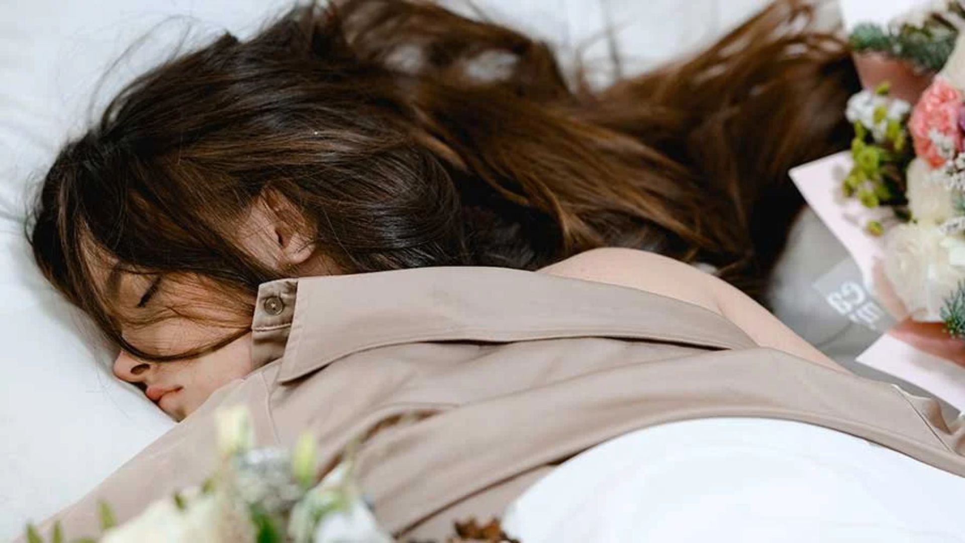 A woman with long brown hair sleeps peacefully on a pillow, surrounded by bouquets of flowers. The tone is serene and calm, conveying rest and tranquility.