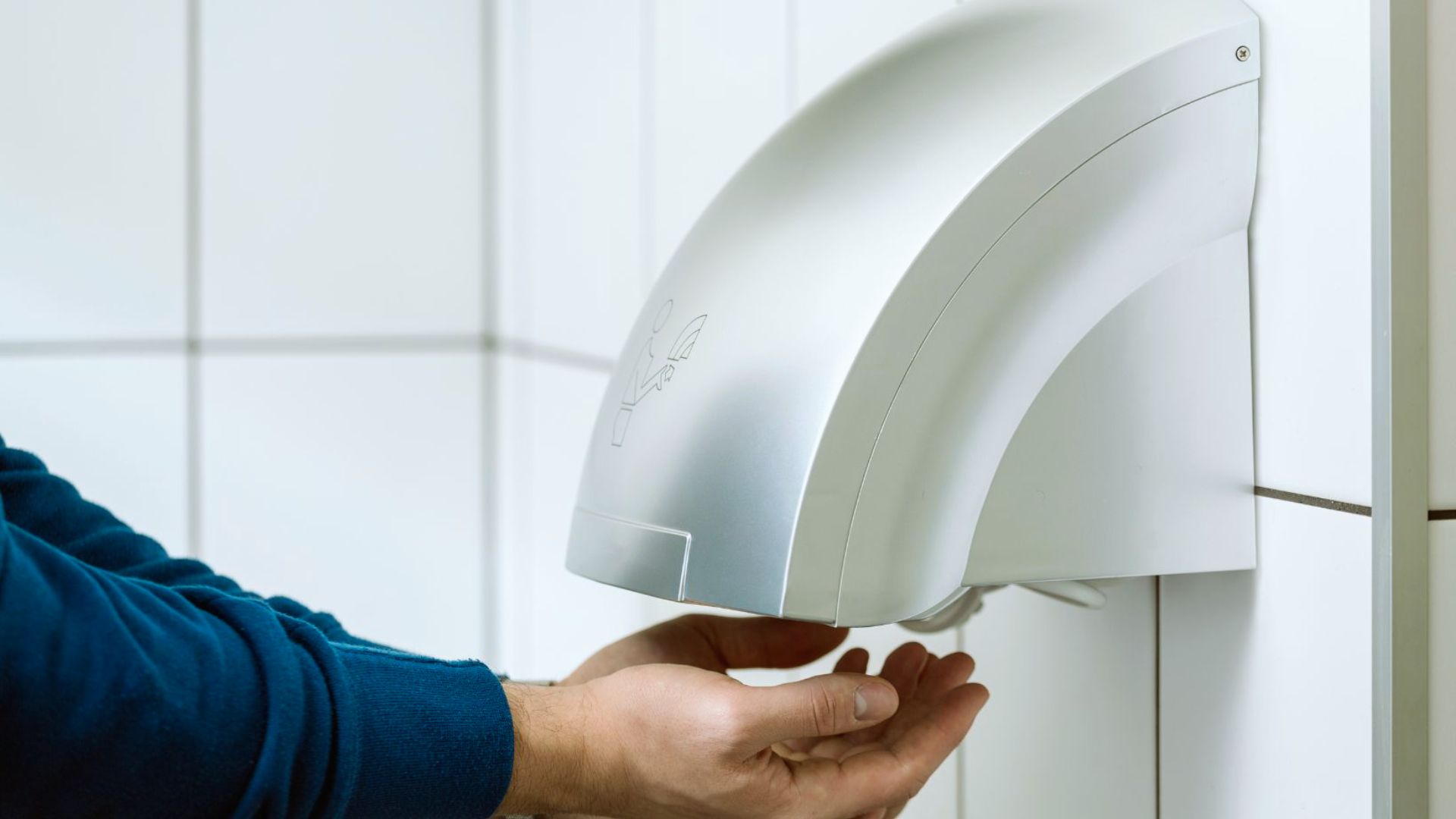 A person dries their hands under an automatic hand dryer mounted on a tiled wall. The setting appears clean and modern, suggesting a public restroom.