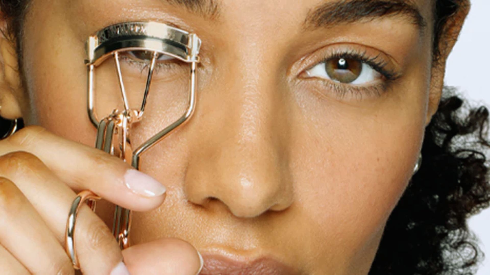 A person uses a gold eyelash curler on their lashes, focusing on one eye. The scene conveys a beauty routine with a calm, focused expression.