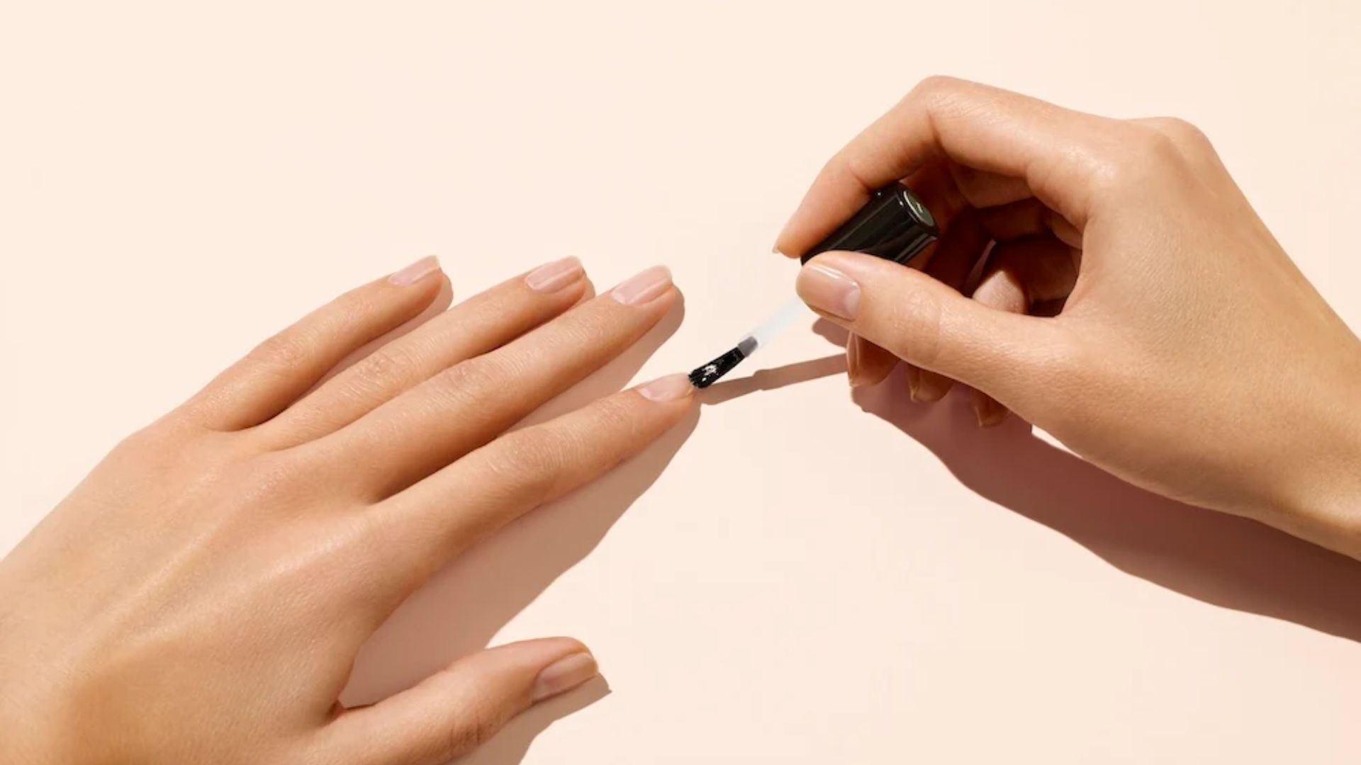 A hand applies clear nail polish to another hand's fingernails on a light peach background. The scene conveys a neat, calm, and self-care atmosphere.
