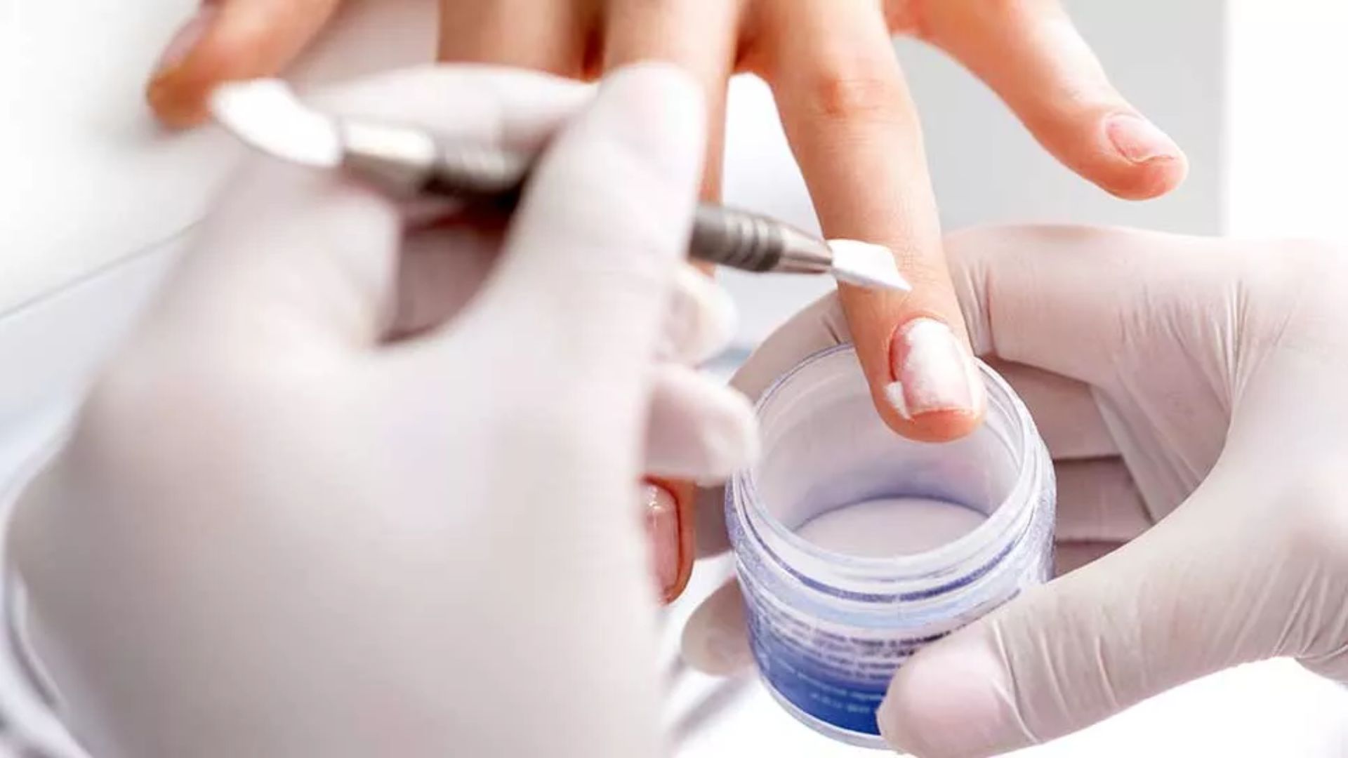 A gloved hand applies white gel to a person's fingernail using a small brush, with an open jar of nail gel nearby, suggesting a manicure session.