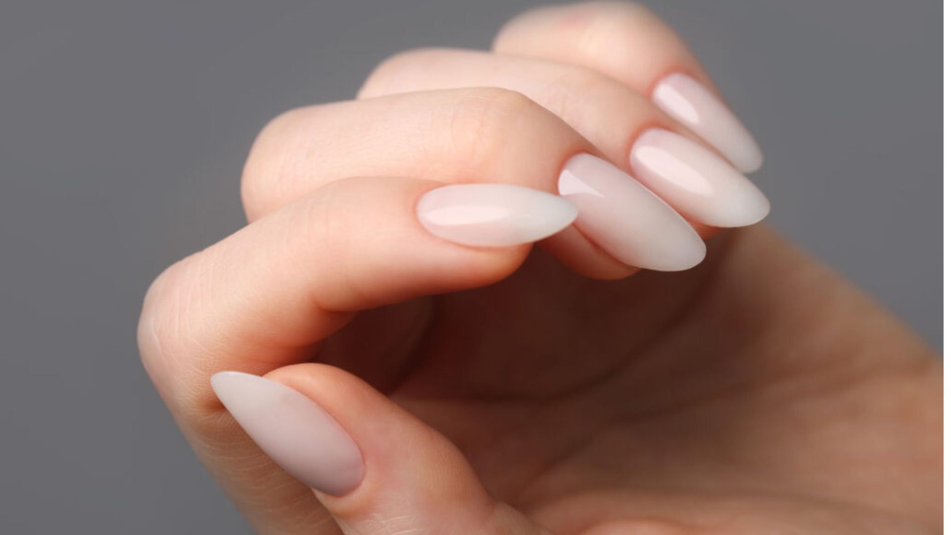 Close-up of a hand with long, almond-shaped nails painted in a smooth, pale pink polish. The background is a neutral gray, creating an elegant and sophisticated tone.