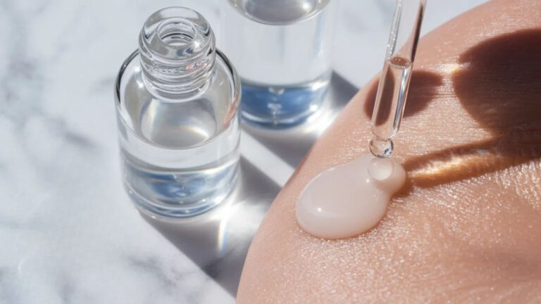 Close-up of a dropper applying serum on skin, beside two glass bottles on a marble surface. The scene conveys a sense of skincare and self-care.