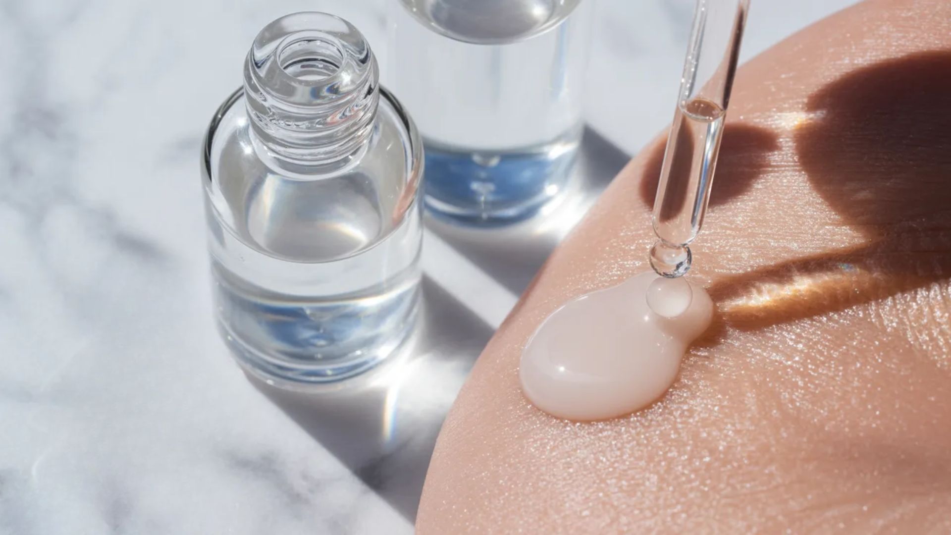 Close-up of a dropper applying serum on skin, beside two glass bottles on a marble surface. The scene conveys a sense of skincare and self-care.