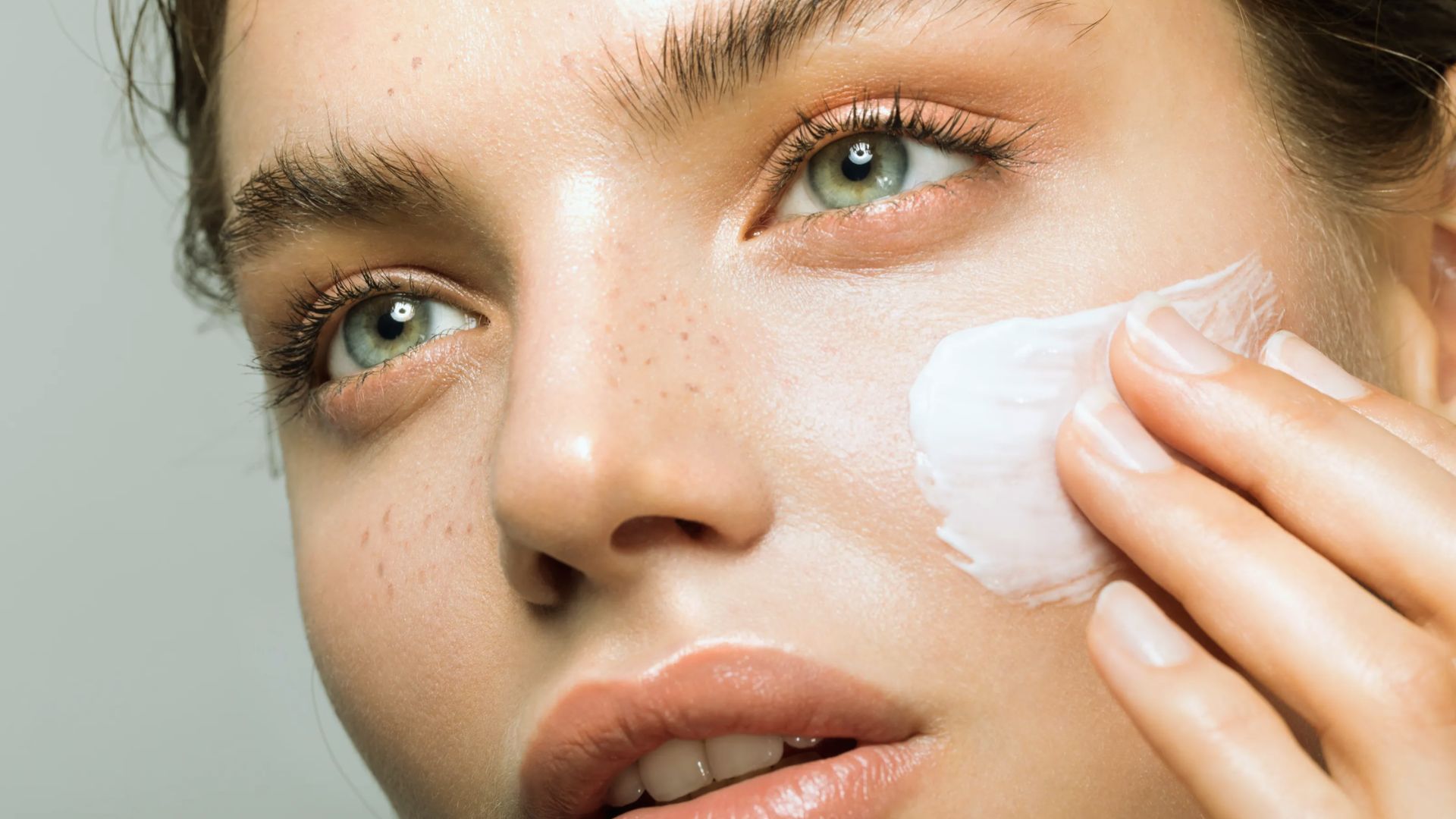 Close-up of a person applying white cream to their cheek. The skin appears smooth and well-hydrated, conveying a sense of care and tranquility.
