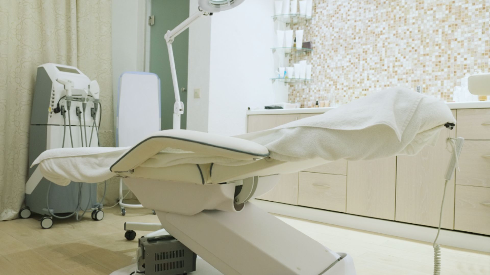 Modern aesthetic clinic room with a sleek treatment chair draped in white towels. Background features a mosaic tiled wall, equipment, and wooden cabinets.