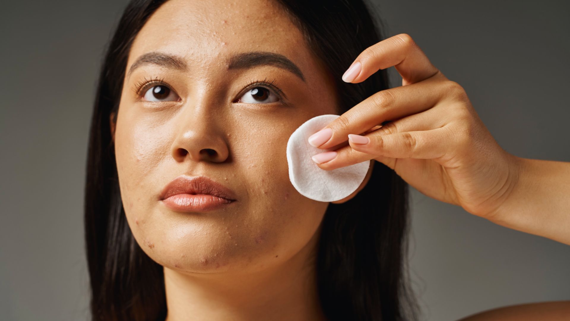 A woman gently applies a cotton pad to her cheek, suggesting a skincare routine. Her expression is calm, and the lighting highlights her natural complexion.