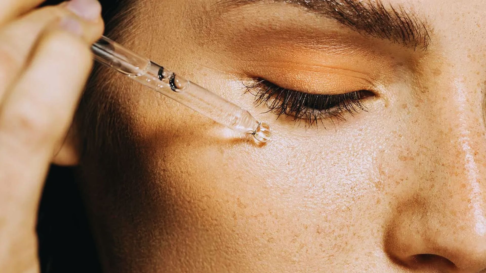 Close-up of a person applying serum under their eye with a dropper. The person has freckles and wears orange eyeshadow, conveying a skincare routine.