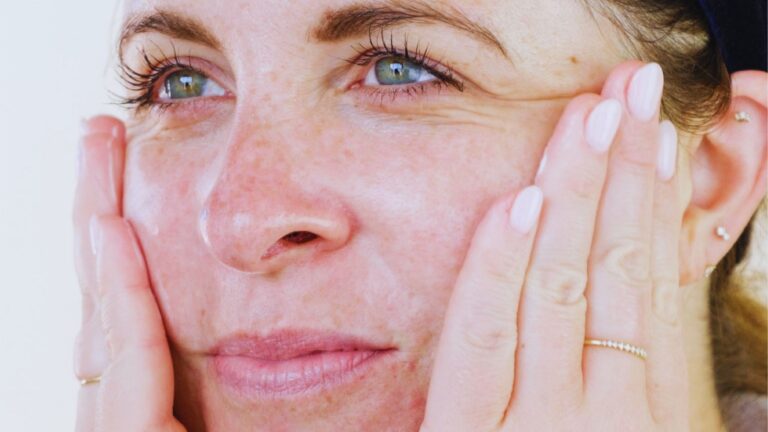 A close-up of a person with light skin gently touching their cheeks, showing freckles and a serene expression. They wear rings and have neatly manicured nails.