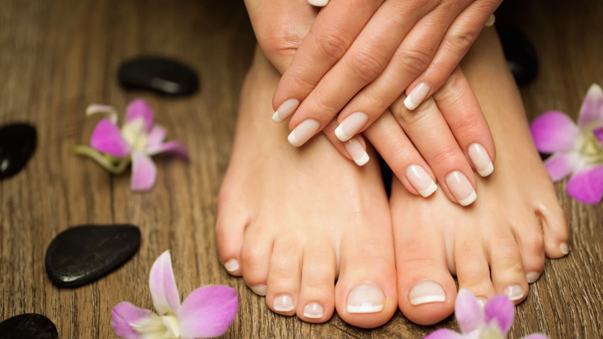 Hands and feet with French manicures and pedicures rest on a wooden surface. Surrounding them are smooth black stones and scattered purple flowers, conveying relaxation.