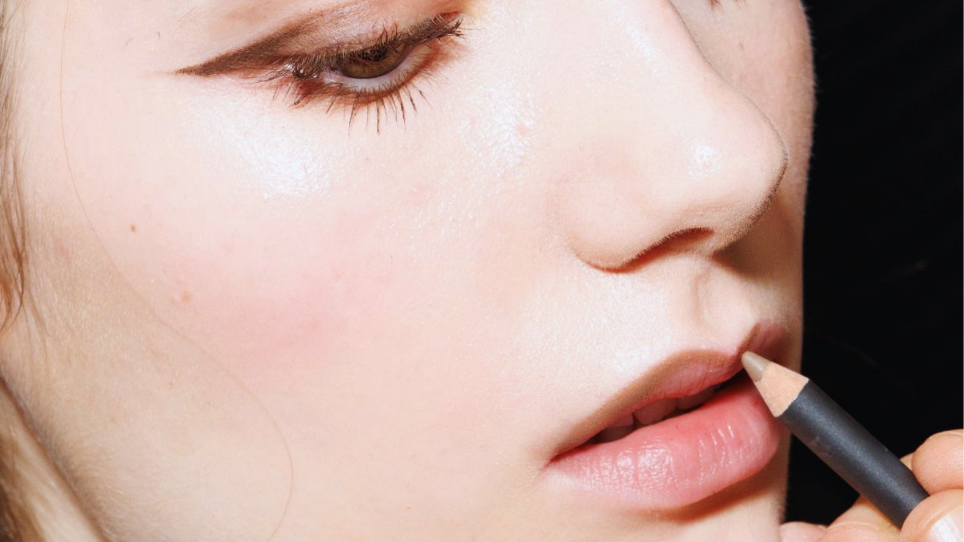 Close-up of a person's face showing a makeup artist applying natural-colored lip liner on their lips. The look is soft, fresh, and focused on precision.