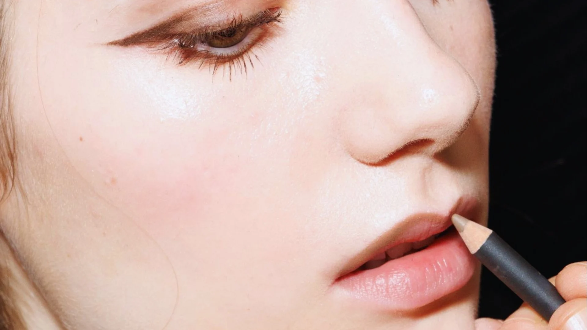 Close-up of a person's face showing a makeup artist applying natural-colored lip liner on their lips. The look is soft, fresh, and focused on precision.