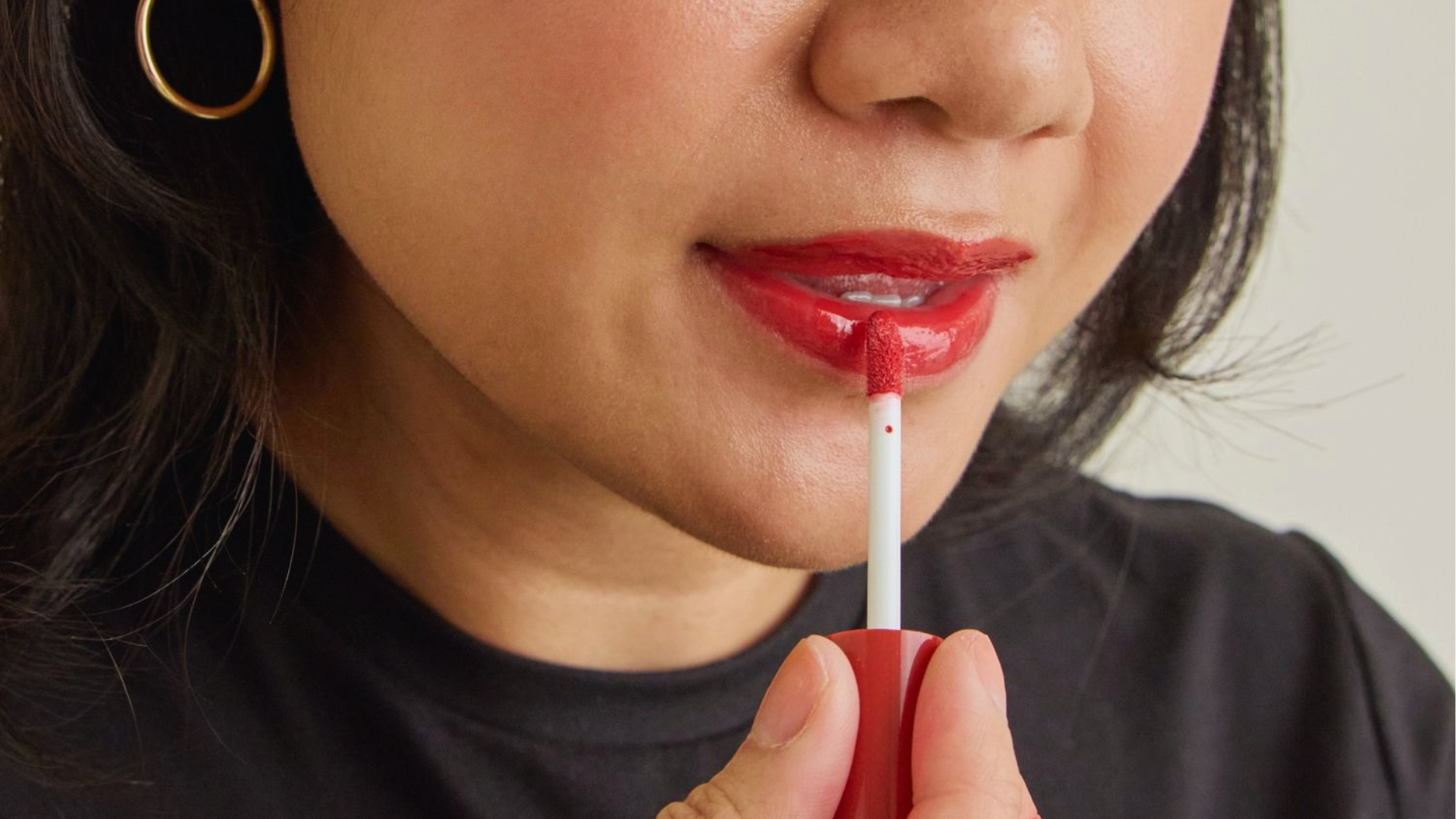 A person applies vibrant red lipstick with a small brush, focusing on precision. They wear a black top and gold hoop earring, creating a polished look.
