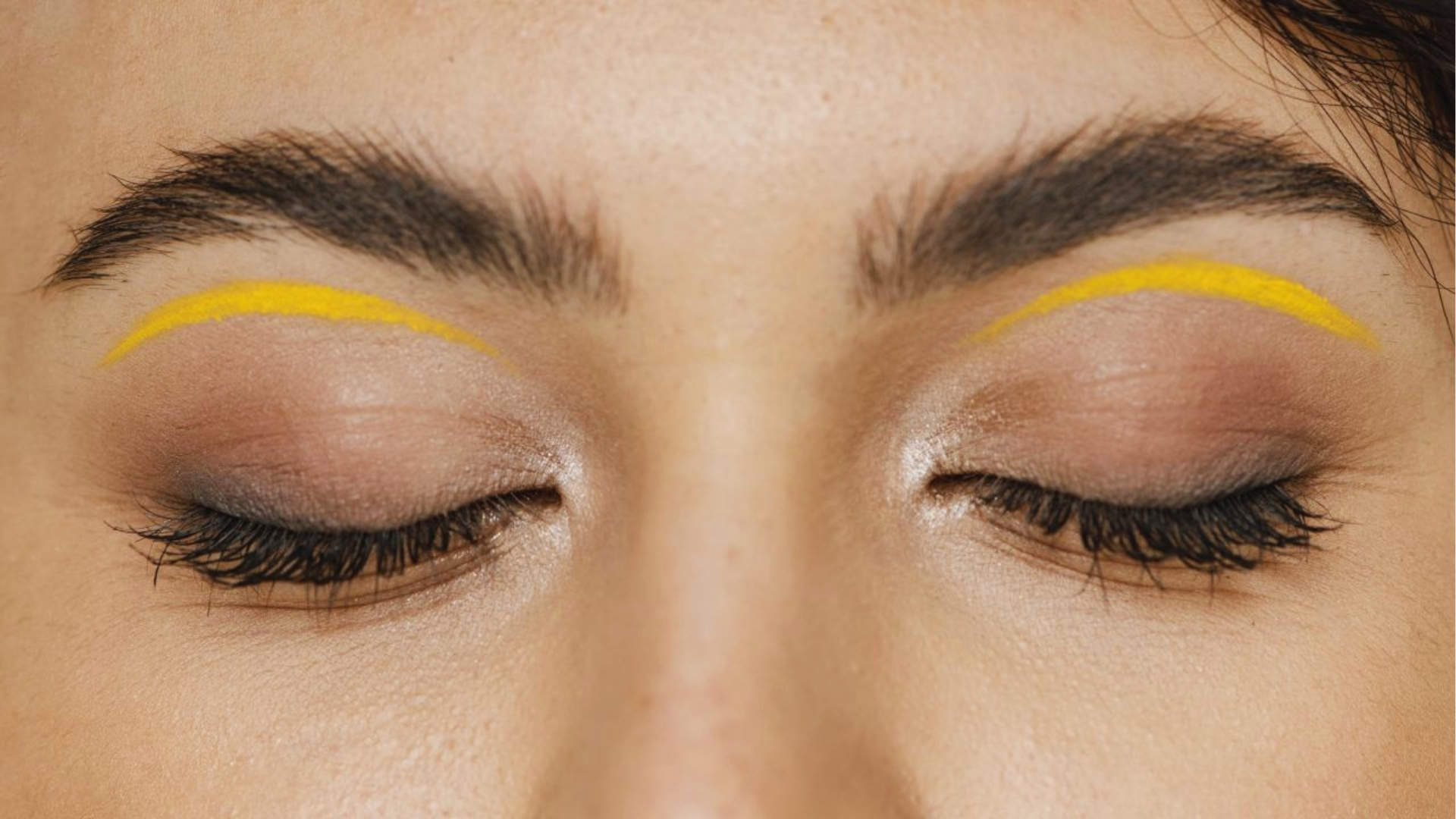 Close-up of a person's closed eyes with bold, thin yellow eyeliner above the lids. The eyebrows are natural, and the skin tone is smooth. The image conveys a creative and stylish makeup look.