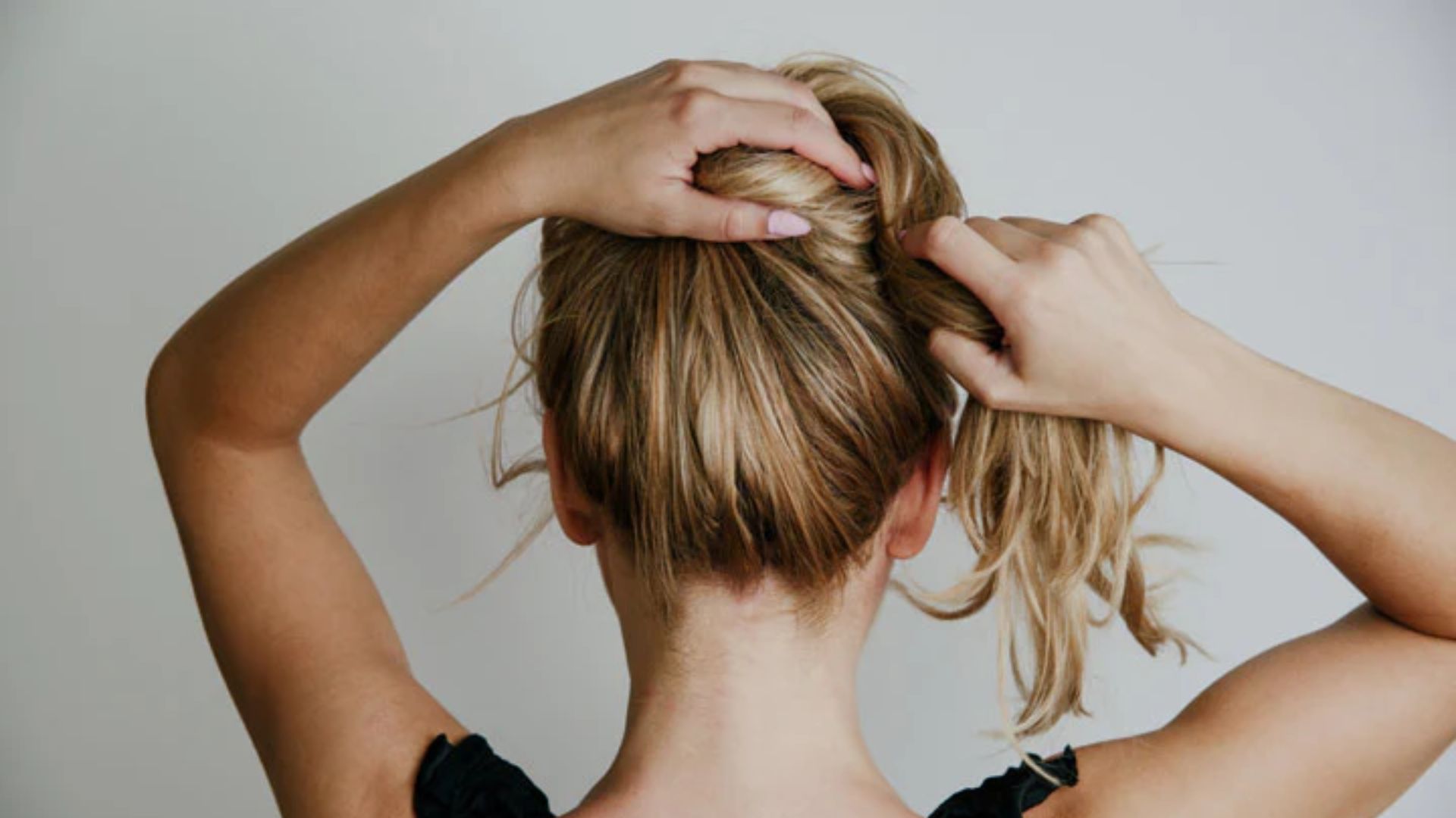 A person with blonde hair is tying their hair into a ponytail, viewed from behind. They wear a black top against a plain, light background.