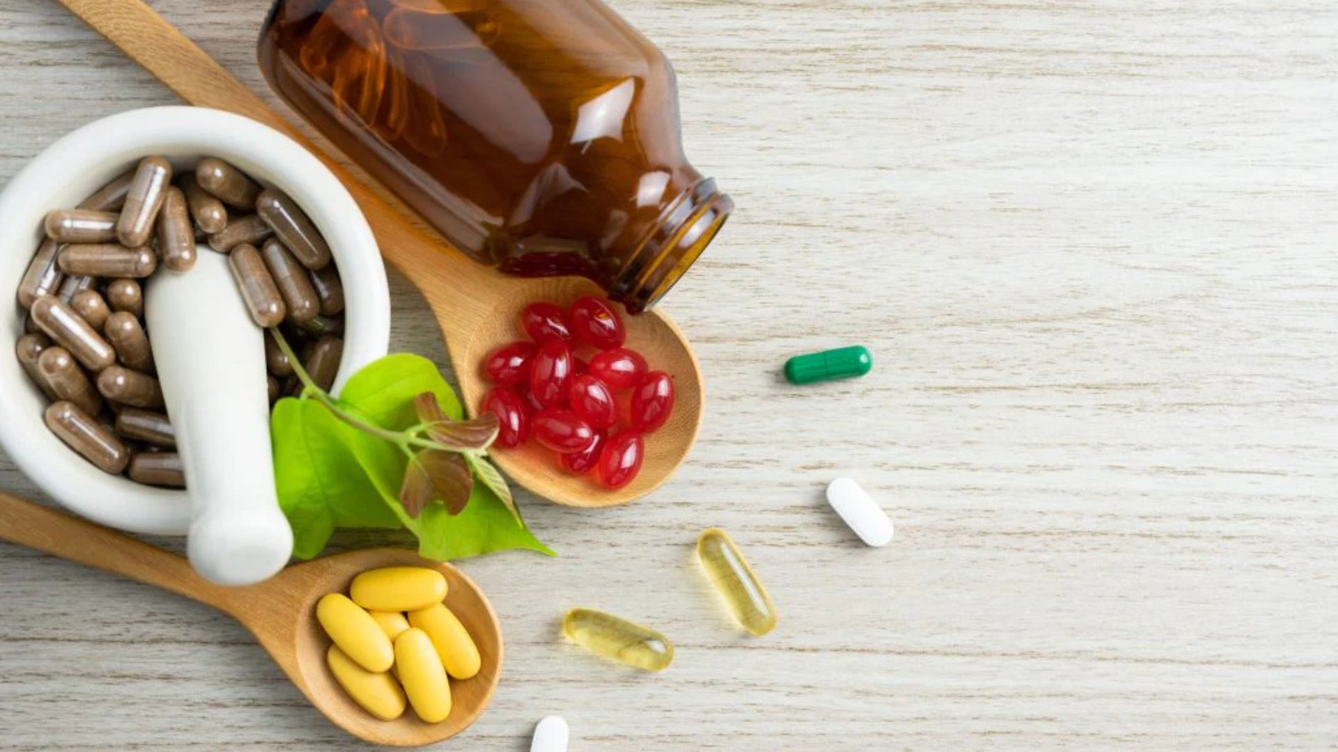 Various pills and capsules scattered on a light wooden surface. A brown bottle, mortar, and pestle are nearby, conveying a health and wellness theme.