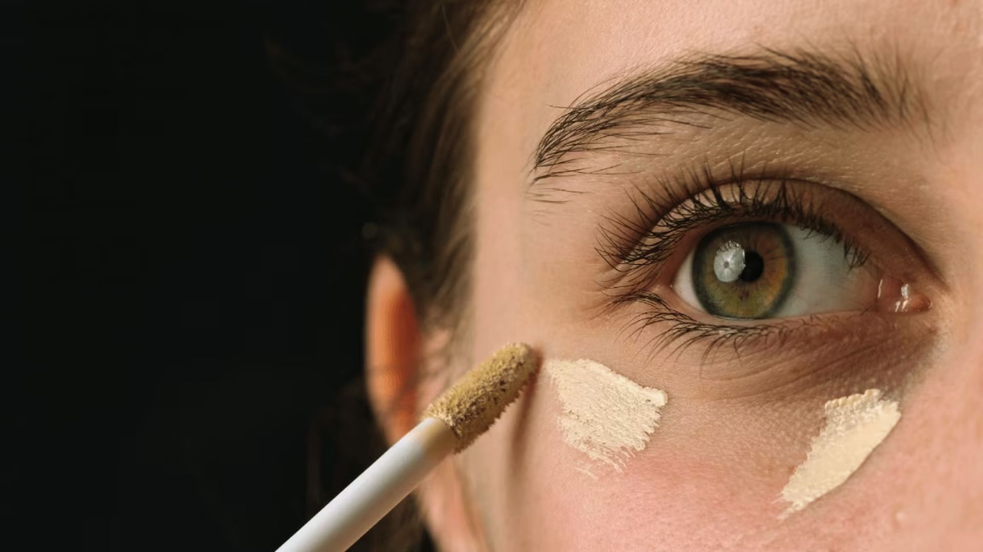 Close-up of a person applying concealer under the eye with a small applicator. The skin tone is highlighted against a dark background, evoking a focused, detailed tone.