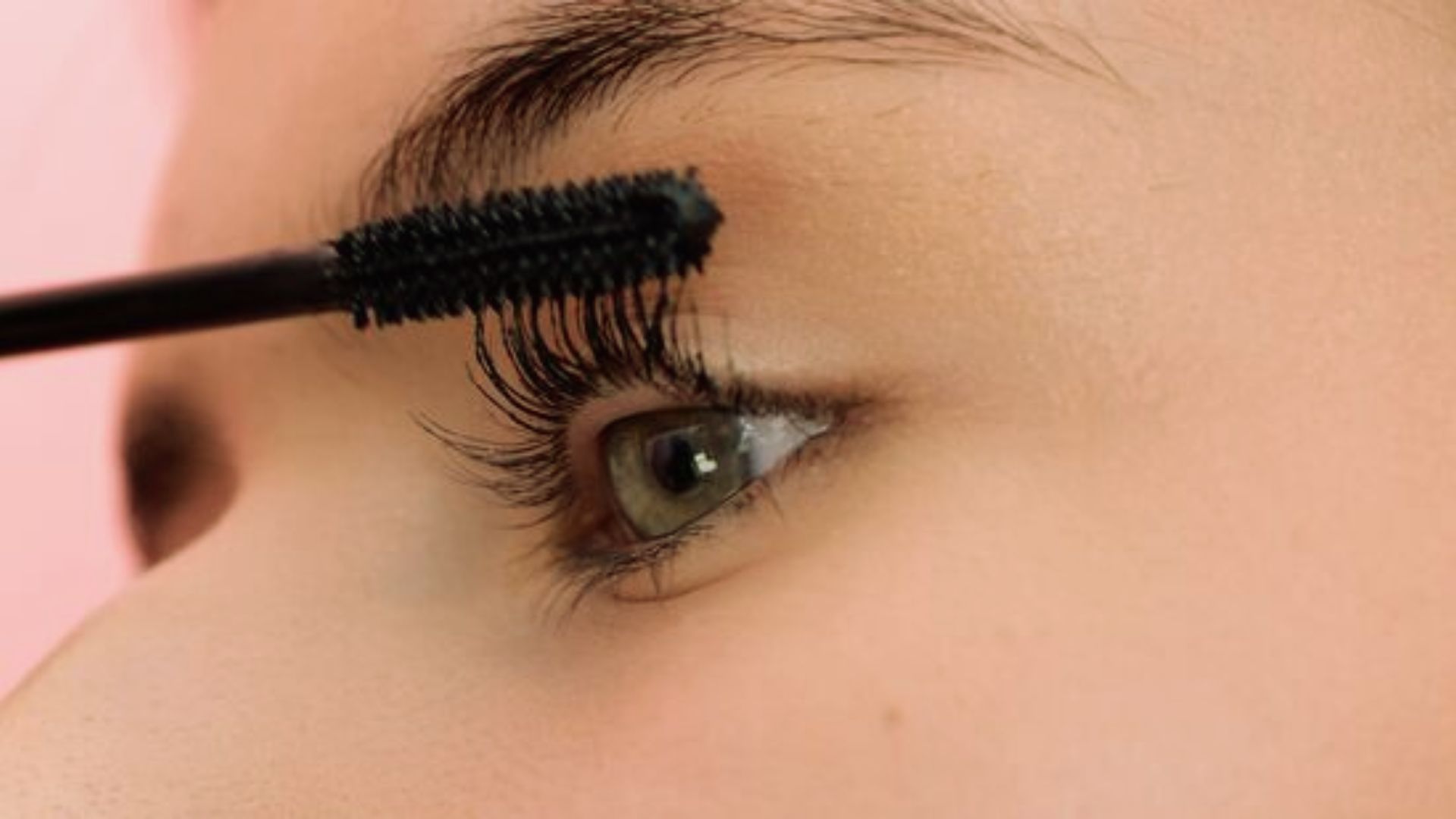 Close-up of a person applying black mascara to their eyelashes. The eye is open, showcasing long lashes and a focused expression against a soft pink background.