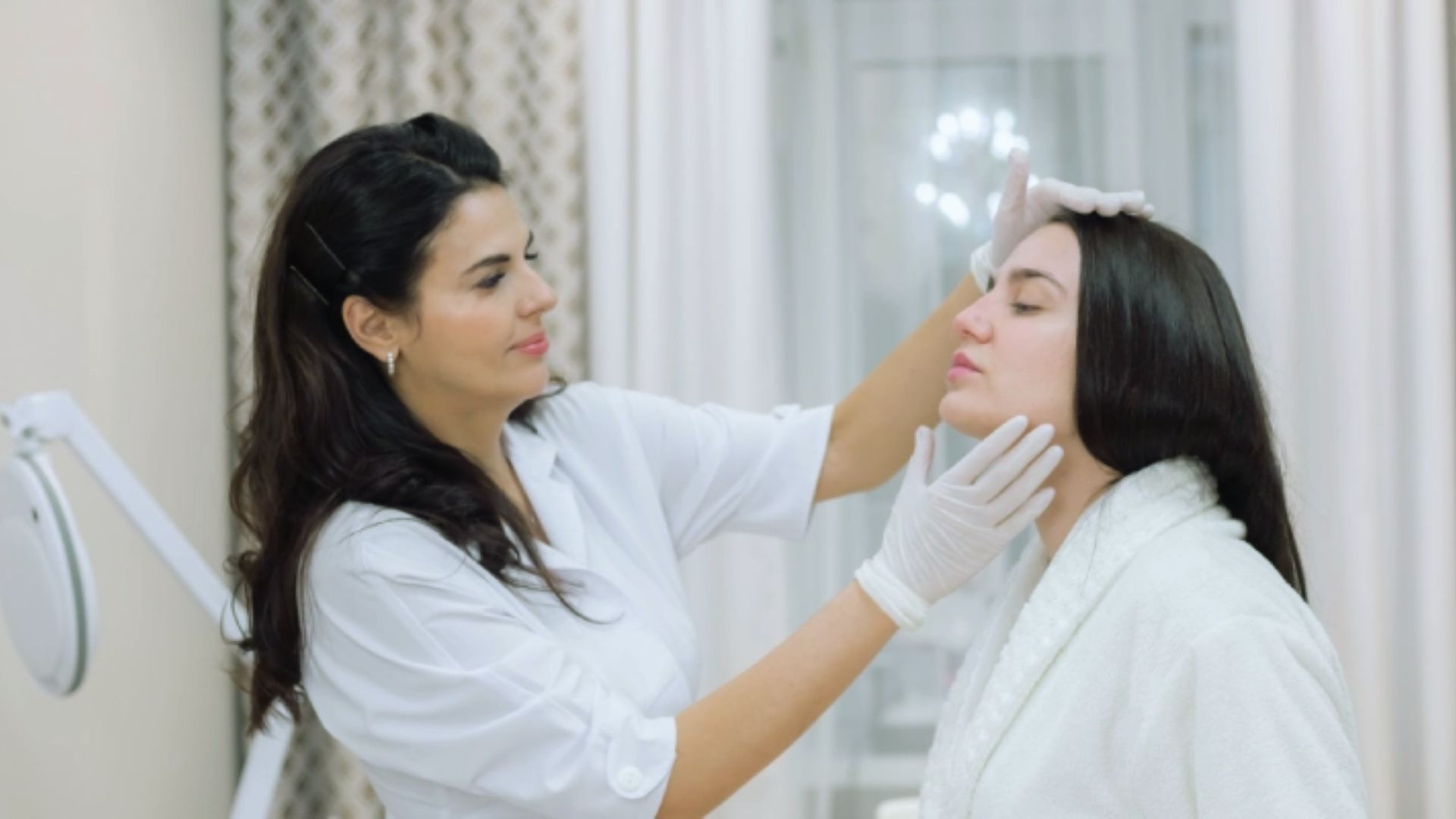 Two women in a white, softly lit room. One, wearing gloves, examines the other's face with care, suggesting a cosmetic or skincare treatment.